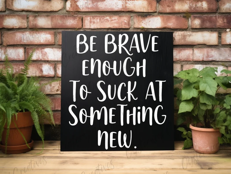 Be brave enough to suck at something new. Black sign with white lettering.